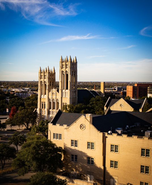 A daytime view of a historic-style church with tall, intricate gothic towers made of light-colored stone. Surrounding the church are smaller buildings with dark roofs, and in the foreground, leafy gre