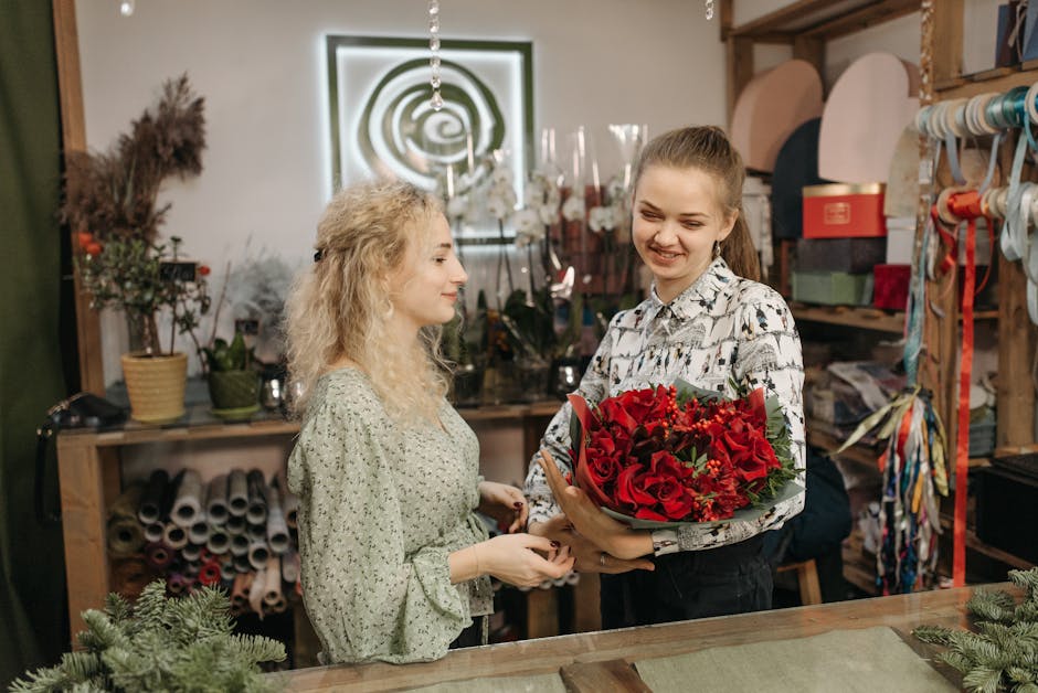 A young woman with light brown hair tied back, wearing a patterned shirt, is smiling as she hands a bouquet of red roses, lilies, and greenery in dark green wrapping to a woman with curly blonde hair 