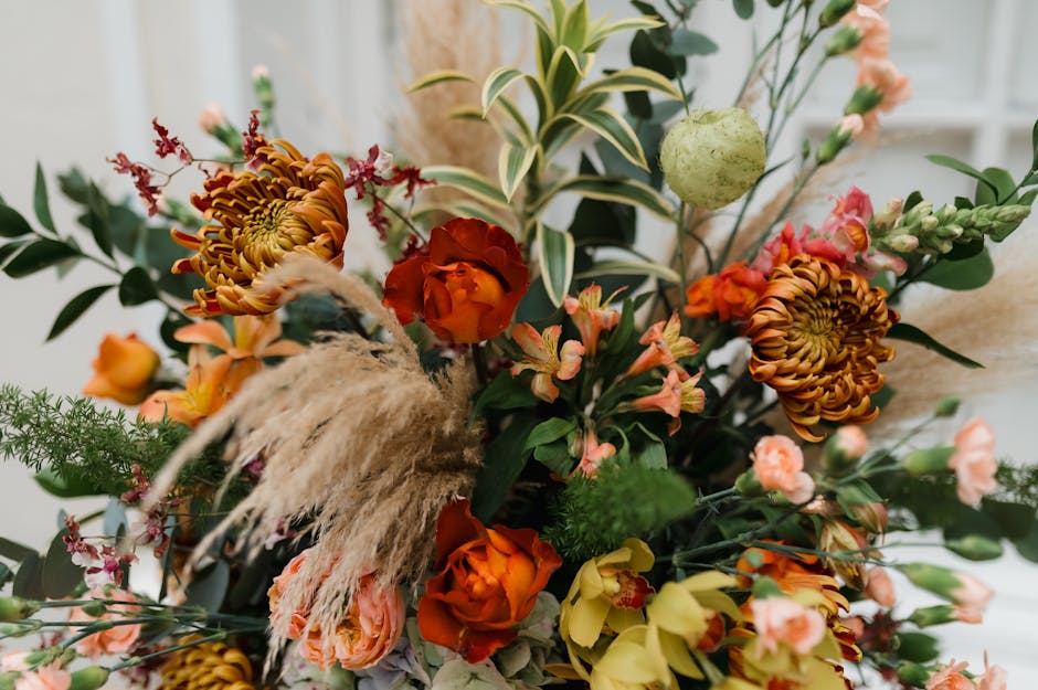 A vibrant floral arrangement featuring a mix of fresh flowers including orange and yellow roses, large brown and yellow dahlias, soft pink and peach spray roses, and delicate pink statice. The bouquet