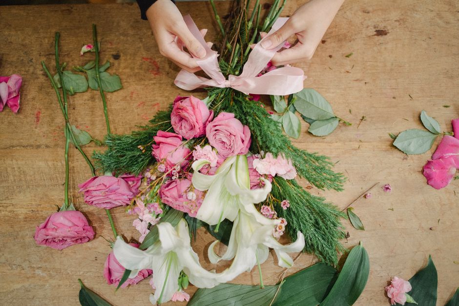A pair of hands arranging a floral bouquet on a wooden table, featuring pink roses, white lilies, and green foliage, with some loose flower petals and stems scattered around. The bouquet is being tied